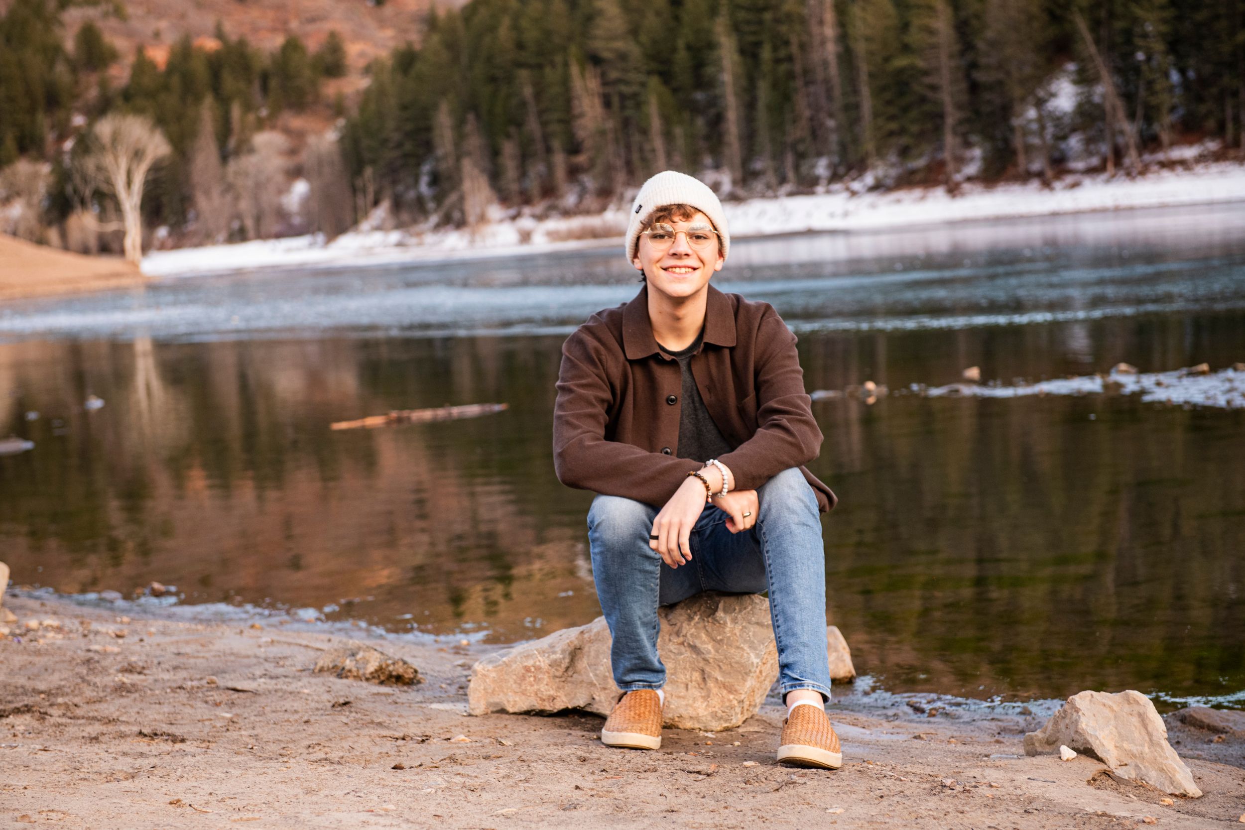 Winter scene at mountain lake with person sitting on rock wearing beanie and brown sweater.