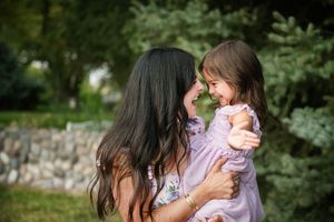 A mother holds her laughing young daughter outdoors, sharing a joyful moment in a lush green garden.