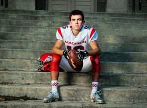 A football player in red and white uniform sits on stone steps holding a football, showcasing athletic gear and equipment.