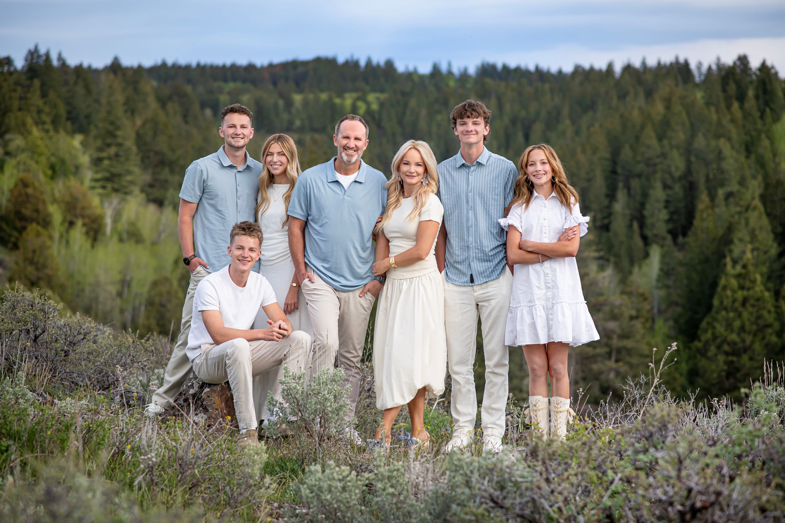 Family of seven posing outdoors in coordinated outfits with evergreen forest backdrop during golden hour.