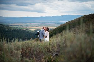 Couple sharing a romantic kiss on a scenic mountain hillside with sweeping valley and mountain range views.