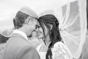 Black and white wedding photo of smiling bride and groom touching foreheads, veil flowing in the wind.