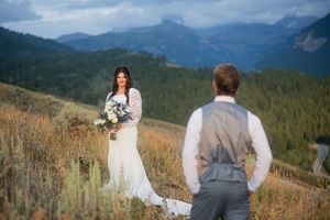 Bride in white lace gown holds bouquet facing groom in gray vest with mountain landscape backdrop.