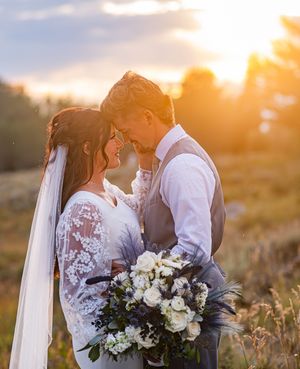 A bride and groom share a romantic forehead touch at golden hour sunset in a field, bride holding white floral bouquet.