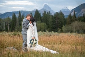 Bride and groom embrace in a mountain meadow, with the Grand Tetons and evergreen trees in the background.