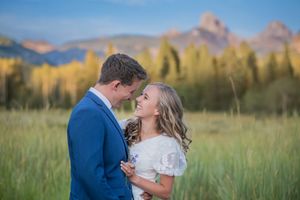 Couple in wedding attire embracing in a mountain meadow with autumn trees and rocky peaks in the background.