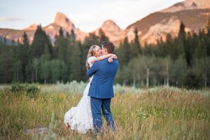 Bride and groom embrace in a mountain meadow at golden hour with rocky peaks glowing in the background.