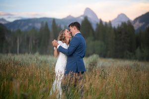 Couple sharing a romantic moment in a mountain meadow at sunset, bride in lace gown and groom in blue suit.