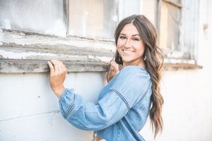 A young woman in a light denim shirt poses casually against a white painted wall while smiling.