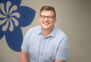 Smiling young man with glasses in a light blue shirt poses in front of a decorative blue floral wall graphic.