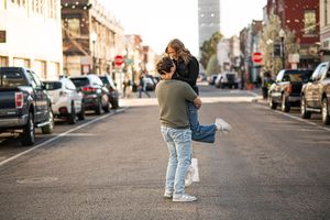 Couple embracing romantically in the middle of a sunny urban street lined with parked cars and shops.
