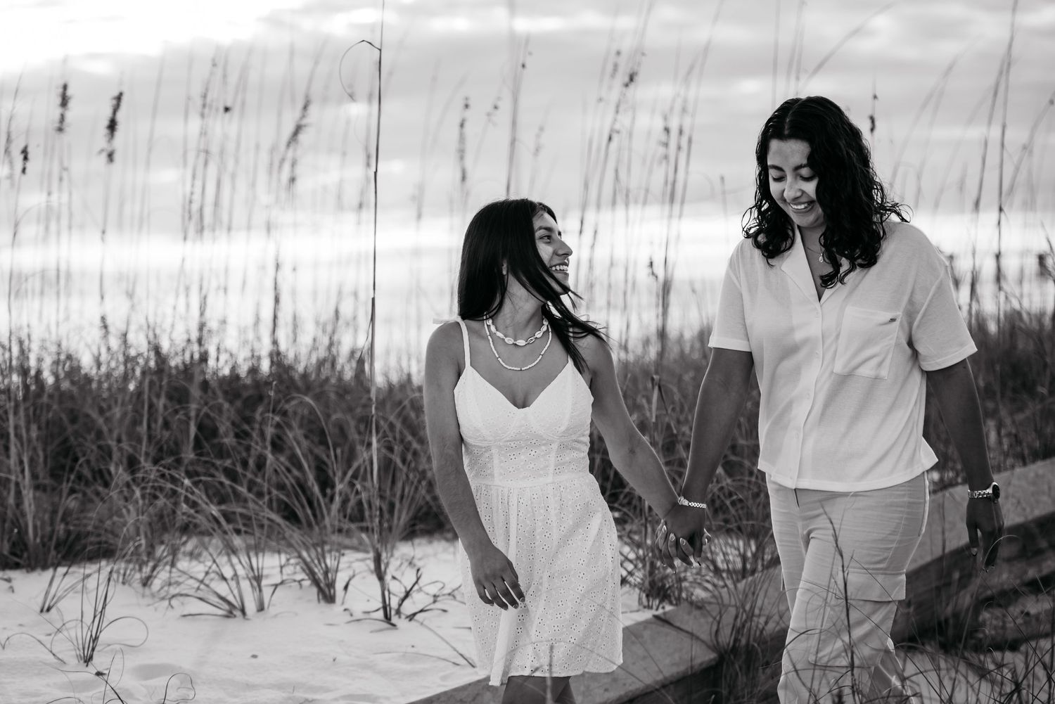 A black and white photo of a couple walking through beach grass at sunset.