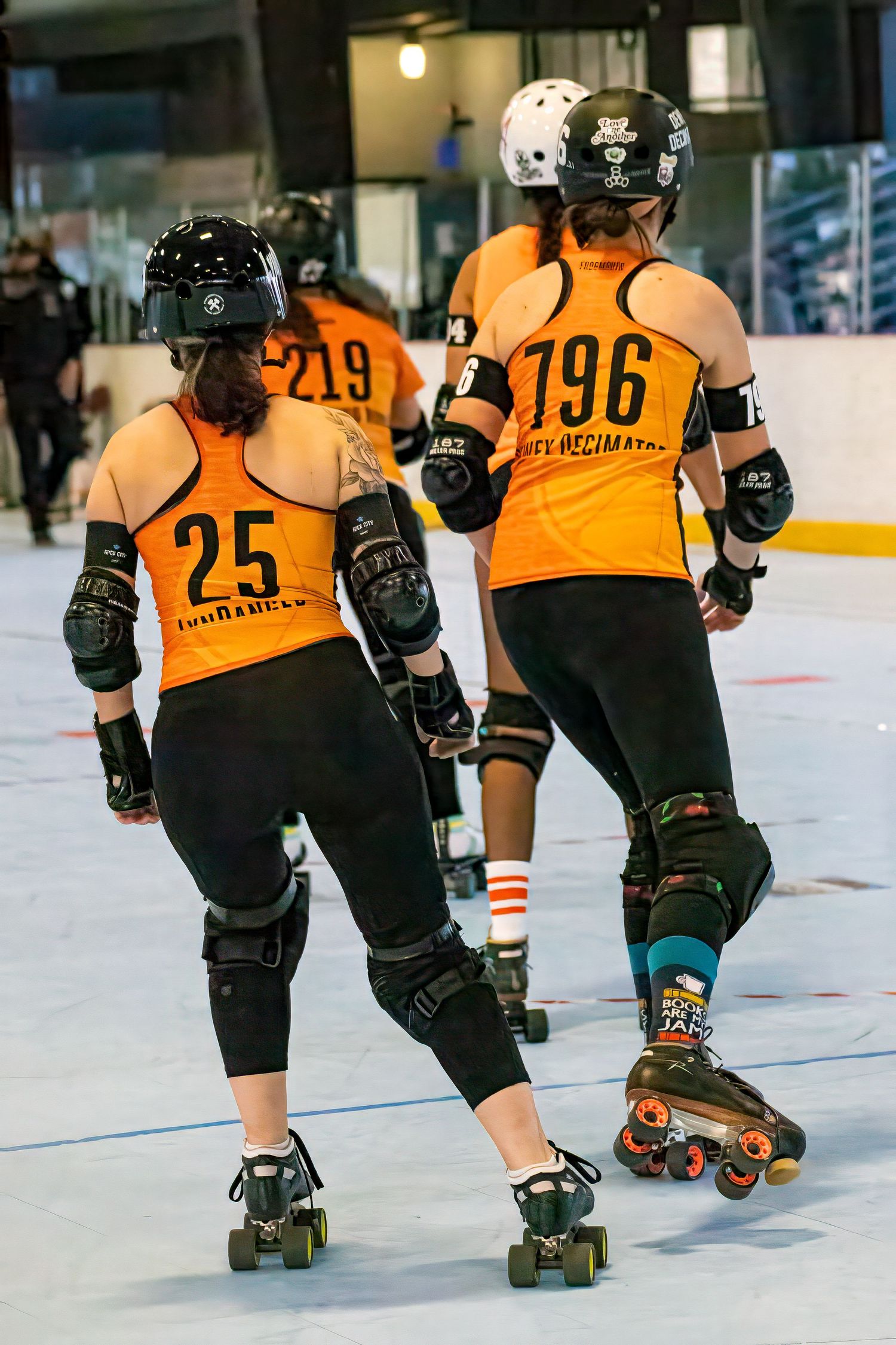 Two roller derby players in orange jerseys numbered 25 and 196 skating on indoor rink wearing protective gear.