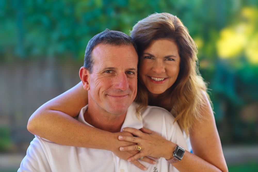 A couple smiling together outdoors on a sunny day with green foliage in the background.