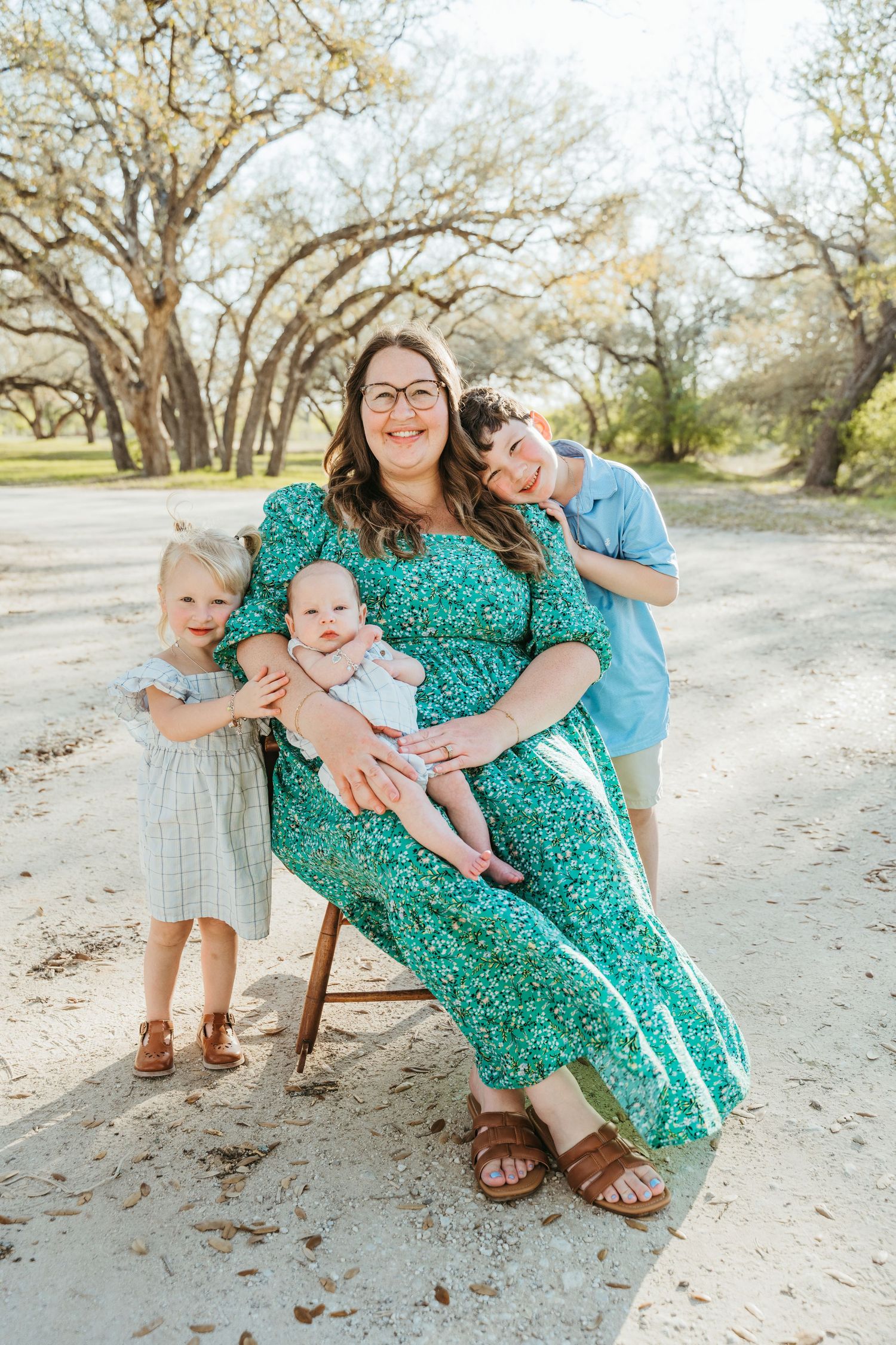 A smiling mother in a green floral dress sits outdoors holding a baby, flanked by a toddler girl and older boy.