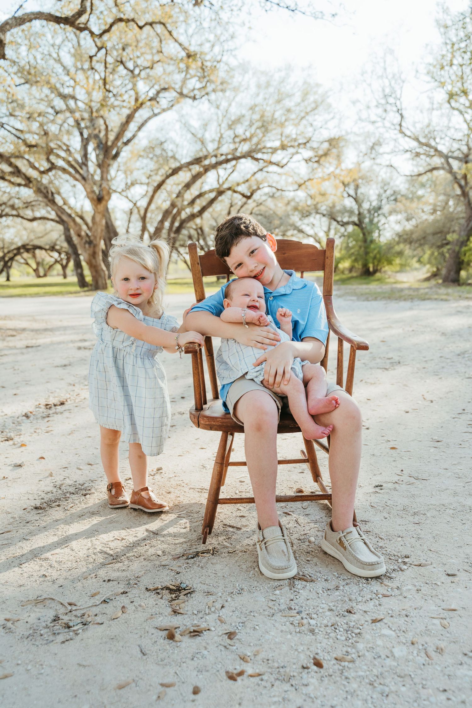 Three siblings pose outdoors, oldest boy sitting in wooden chair holding baby, toddler girl in blue dress standing beside them.