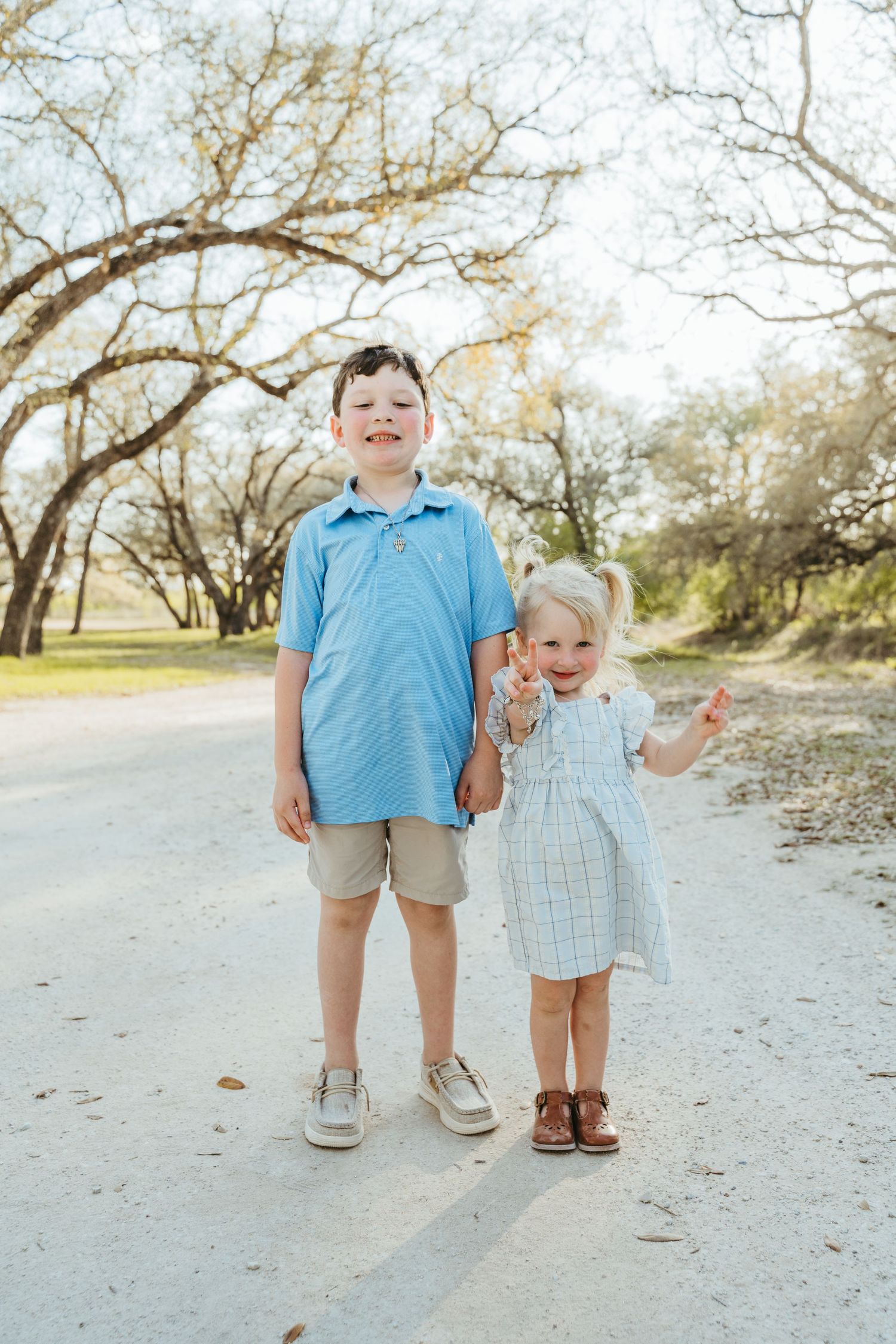 Two children, a boy in a blue polo and a girl in a plaid dress, smile on a sunny dirt path surrounded by trees.
