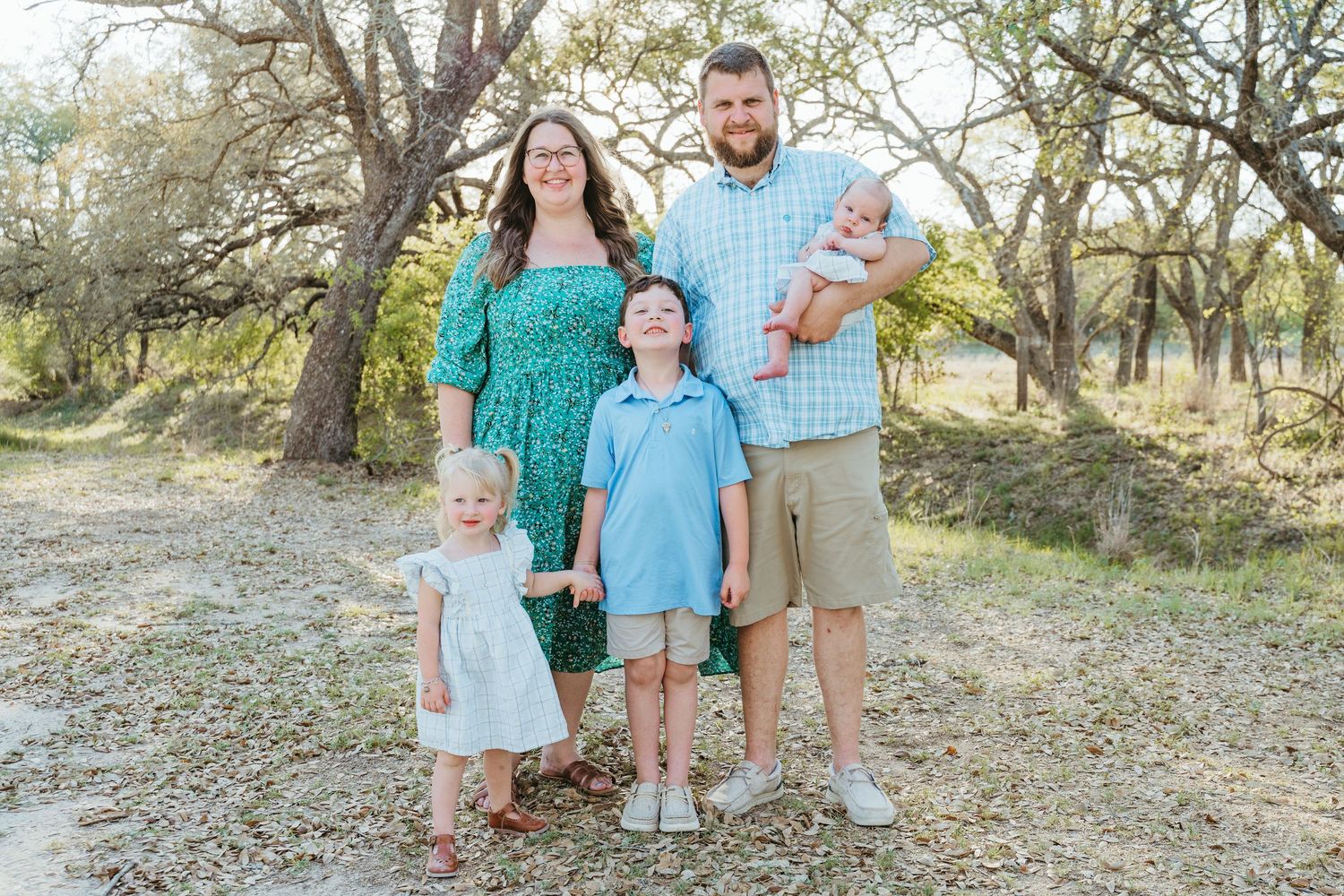 Family of five posing outdoors among trees, parents with three young children dressed in coordinating blue and green outfits.