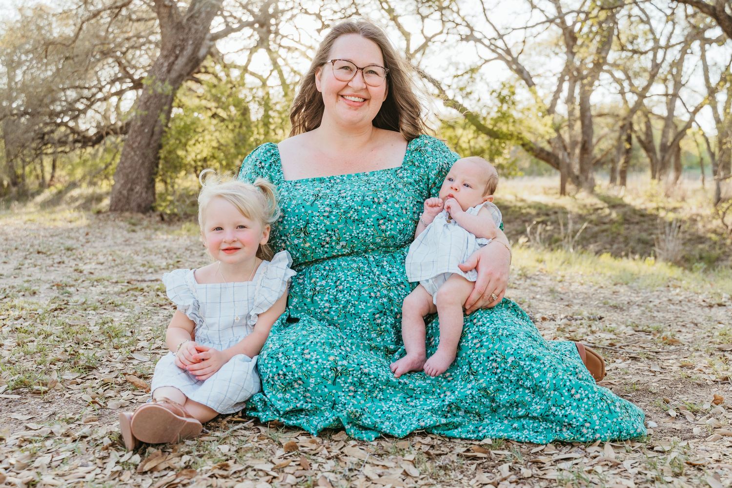 A smiling mother in a green floral dress sits outdoors with her toddler daughter and infant baby in a sunny wooded setting.