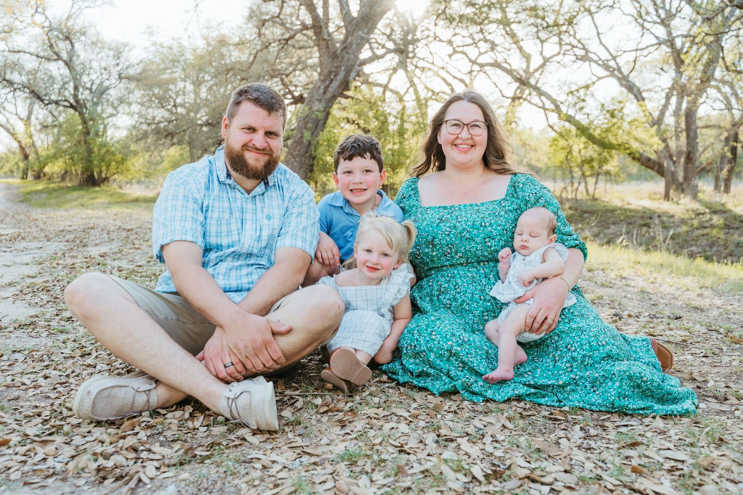 Happy family of five posing outdoors, parents with three young children sitting on ground among trees.