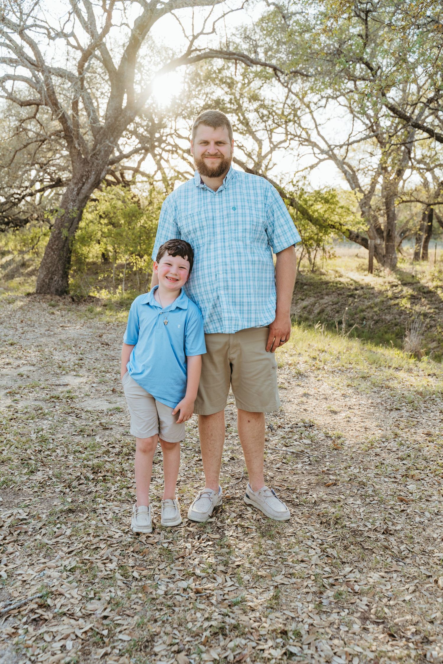 A man and young boy in matching blue shirts and khaki shorts pose outdoors among trees in golden sunlight.