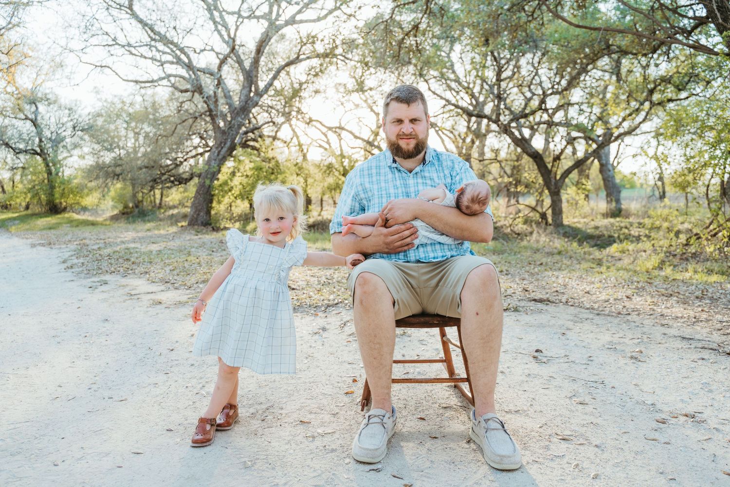 Father sits on stool holding newborn twins outdoors while toddler daughter in white dress stands beside him.