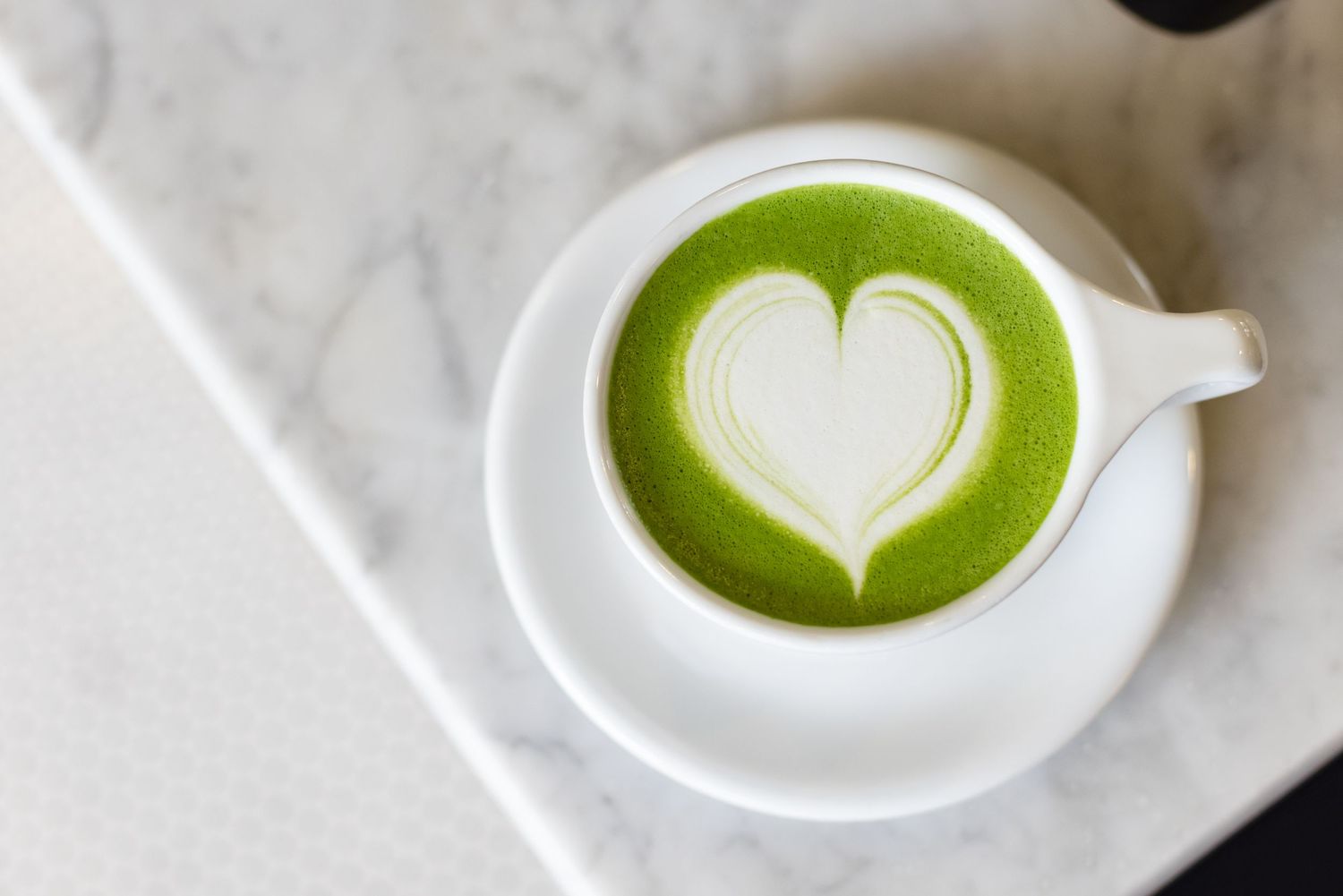 A green matcha latte with white heart-shaped latte art in a white ceramic cup on marble surface.