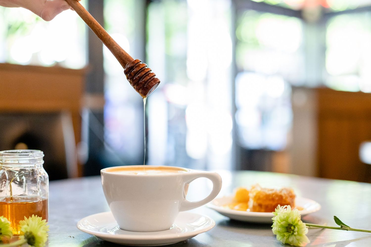 Honey drizzles from a wooden dipper into a white coffee cup alongside a plate with orange garnish and greenery.