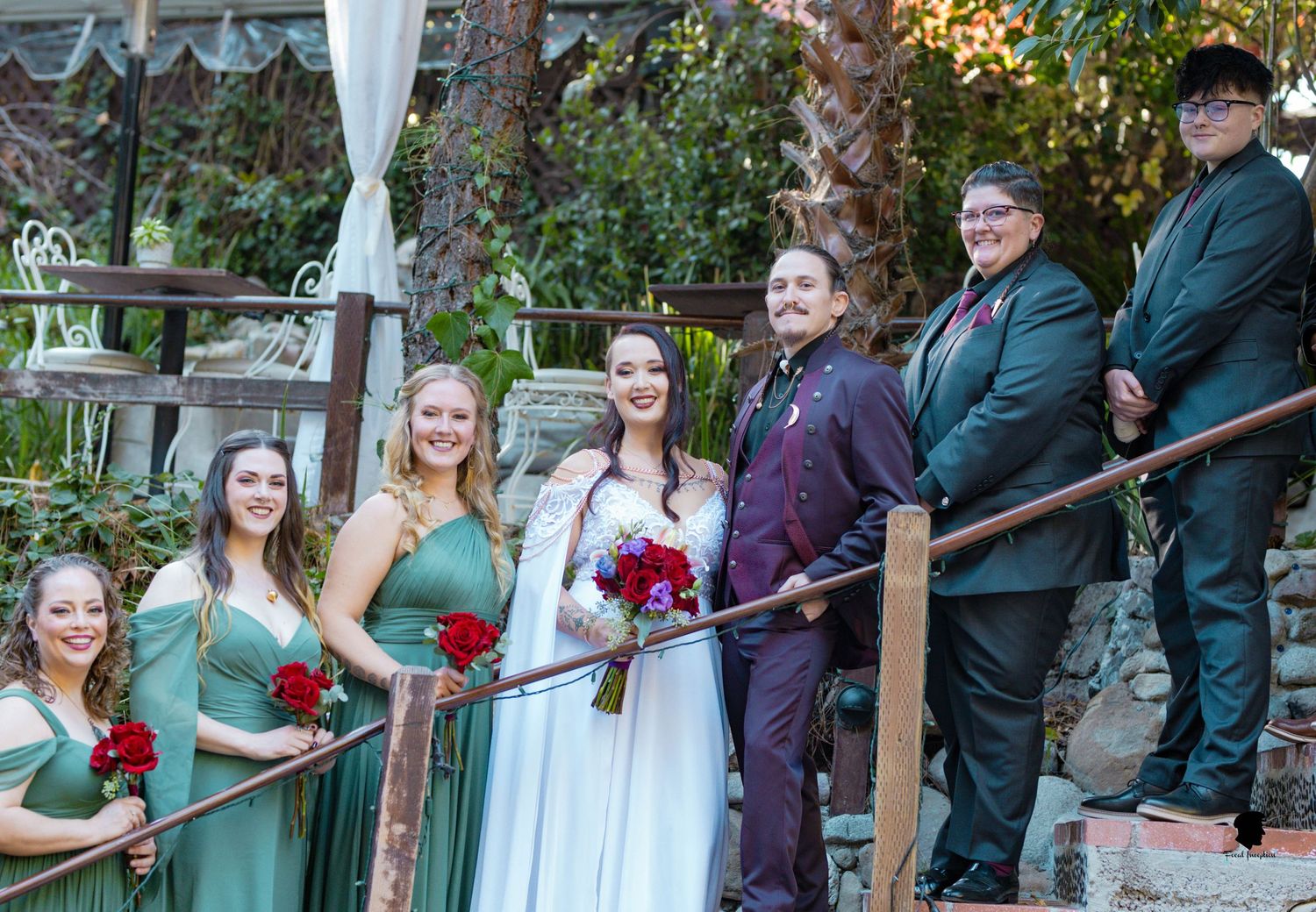 Wedding party in green bridesmaid dresses and formal attire poses on wooden deck.