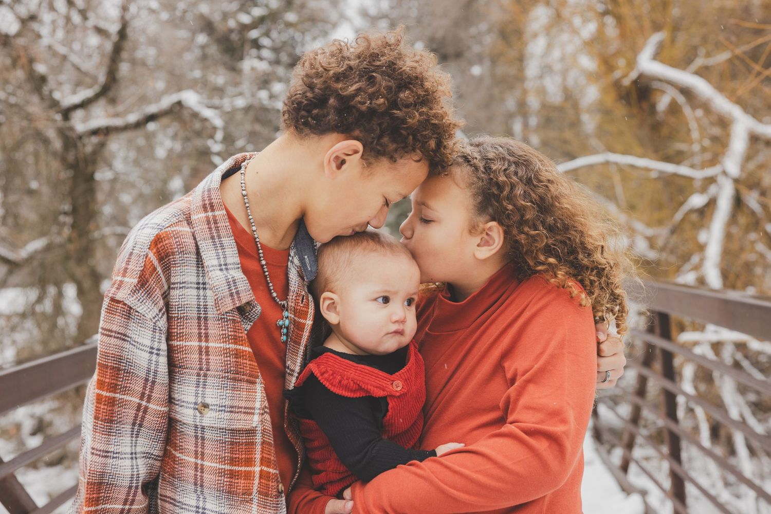 Two young parents with curly hair embrace their infant baby on a snowy bridge in winter.