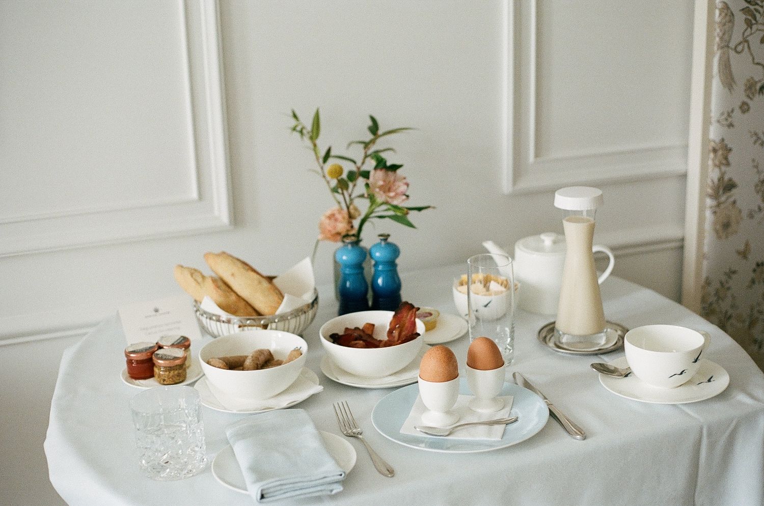 Elegant breakfast table setting with blue vase, fresh flowers, eggs, bread, milk and tea cups on white tablecloth.
