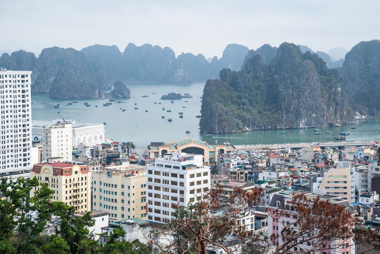 Aerial cityscape of Halong Bay in Vietnam showing urban buildings against dramatic limestone karst formations and boats in water.