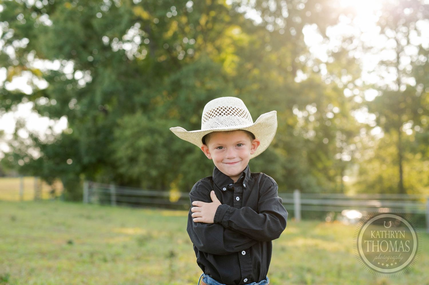 Child in cowboy hat and black shirt stands in sunlit farm field.