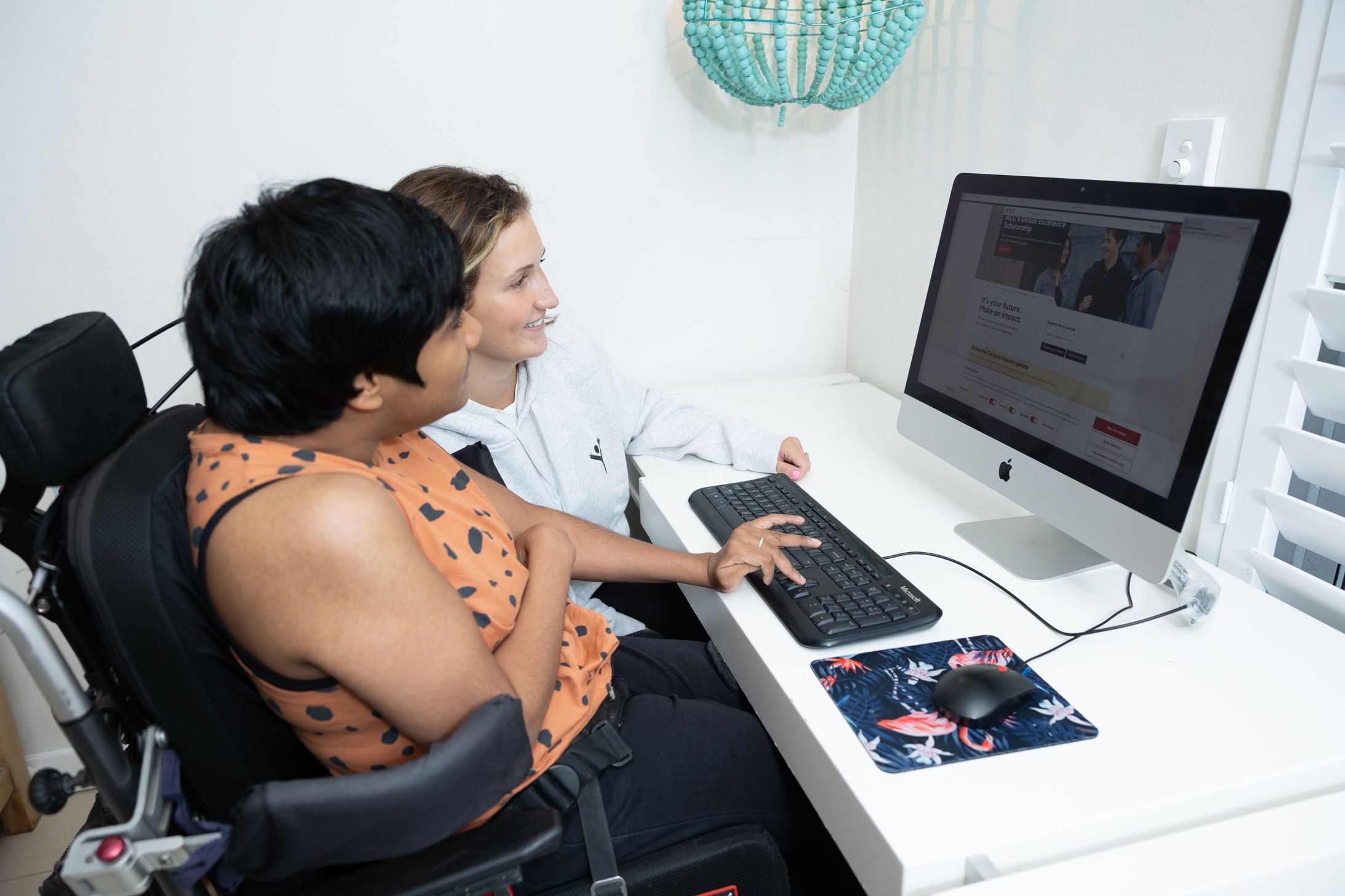Two people share a desk while working together on a computer with a turquoise pendant lamp hanging above.