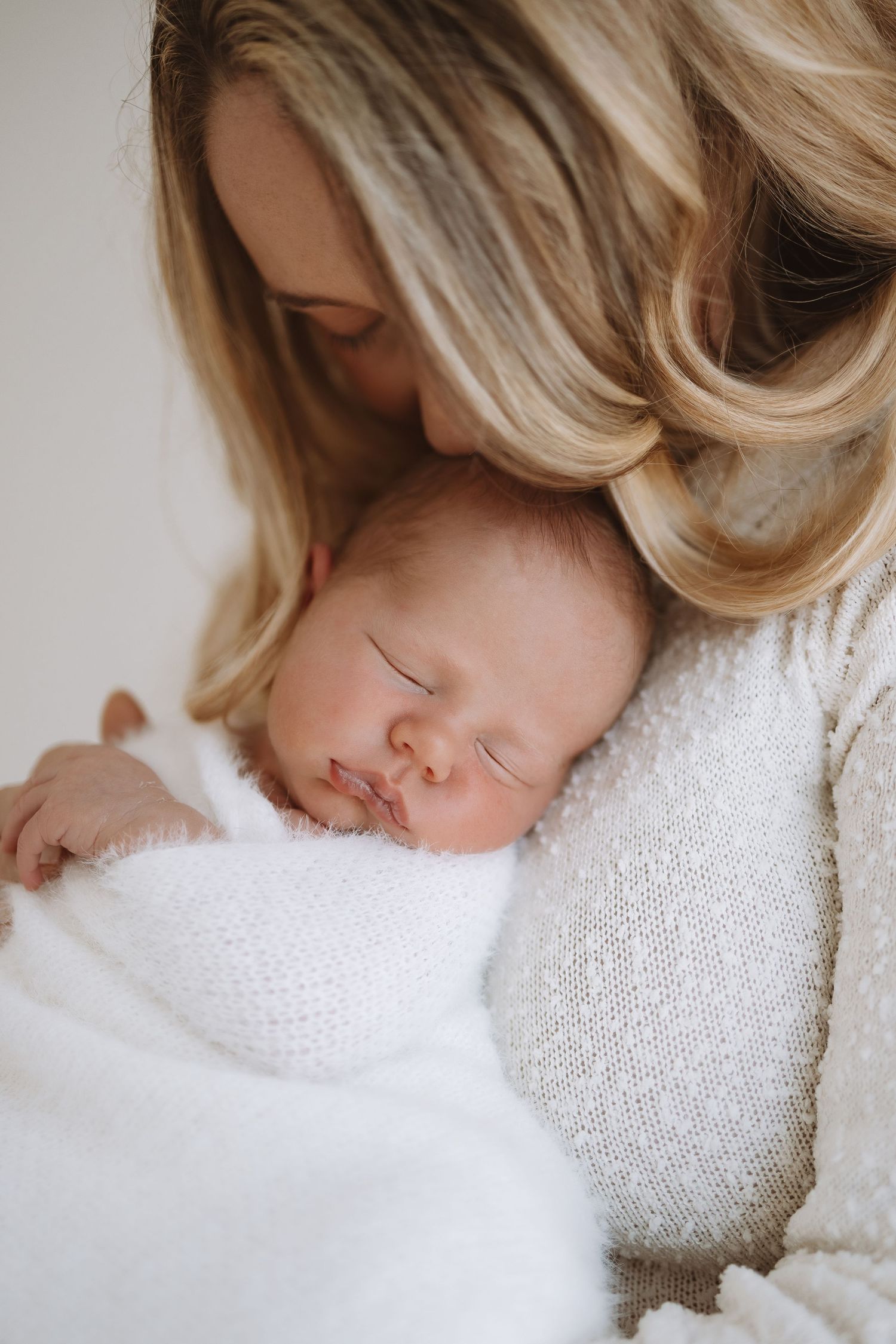 A peacefully sleeping newborn baby lies on white fabric while gentle hands tenderly touch their face.