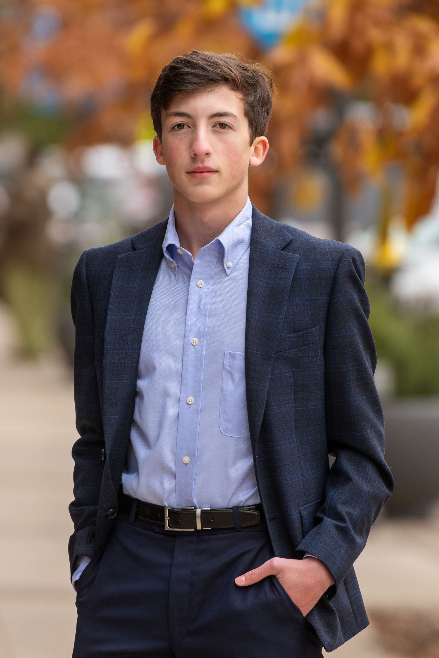 Professional outdoor senior portrait showing young man in a navy suit and light blue button-down shirt against autumn foliage.