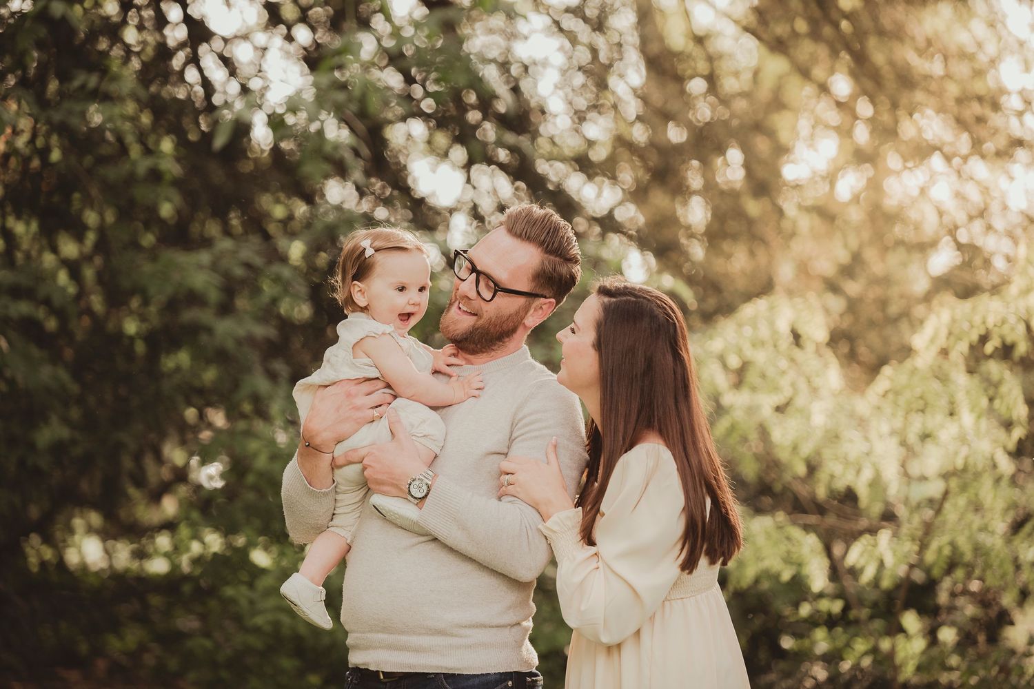 A family in matching cream sweaters embraces outdoors during golden hour in a sunlit garden setting.
