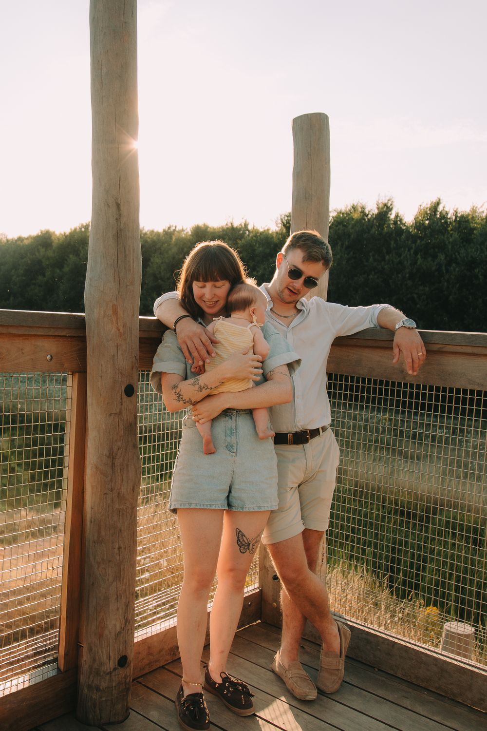 Young family with baby posing on wooden deck at sunset, surrounded by lush greenery.