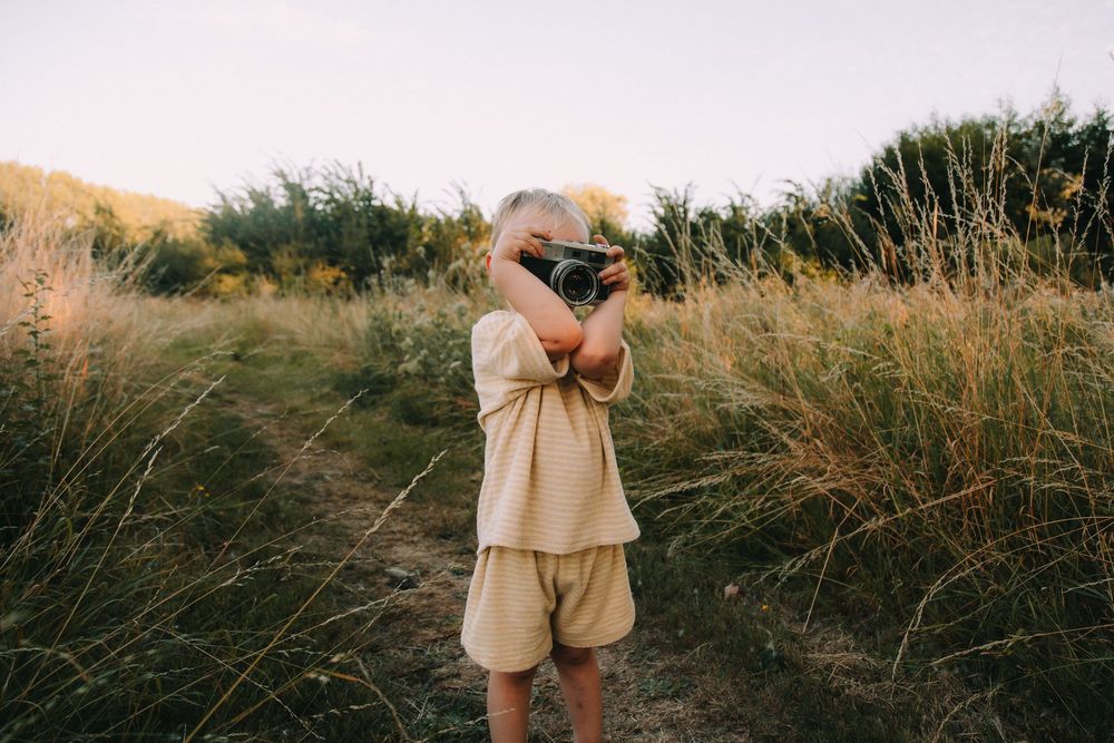 Child in beige outfit photographing nature in a golden field with camera, surrounded by tall grass and greenery.