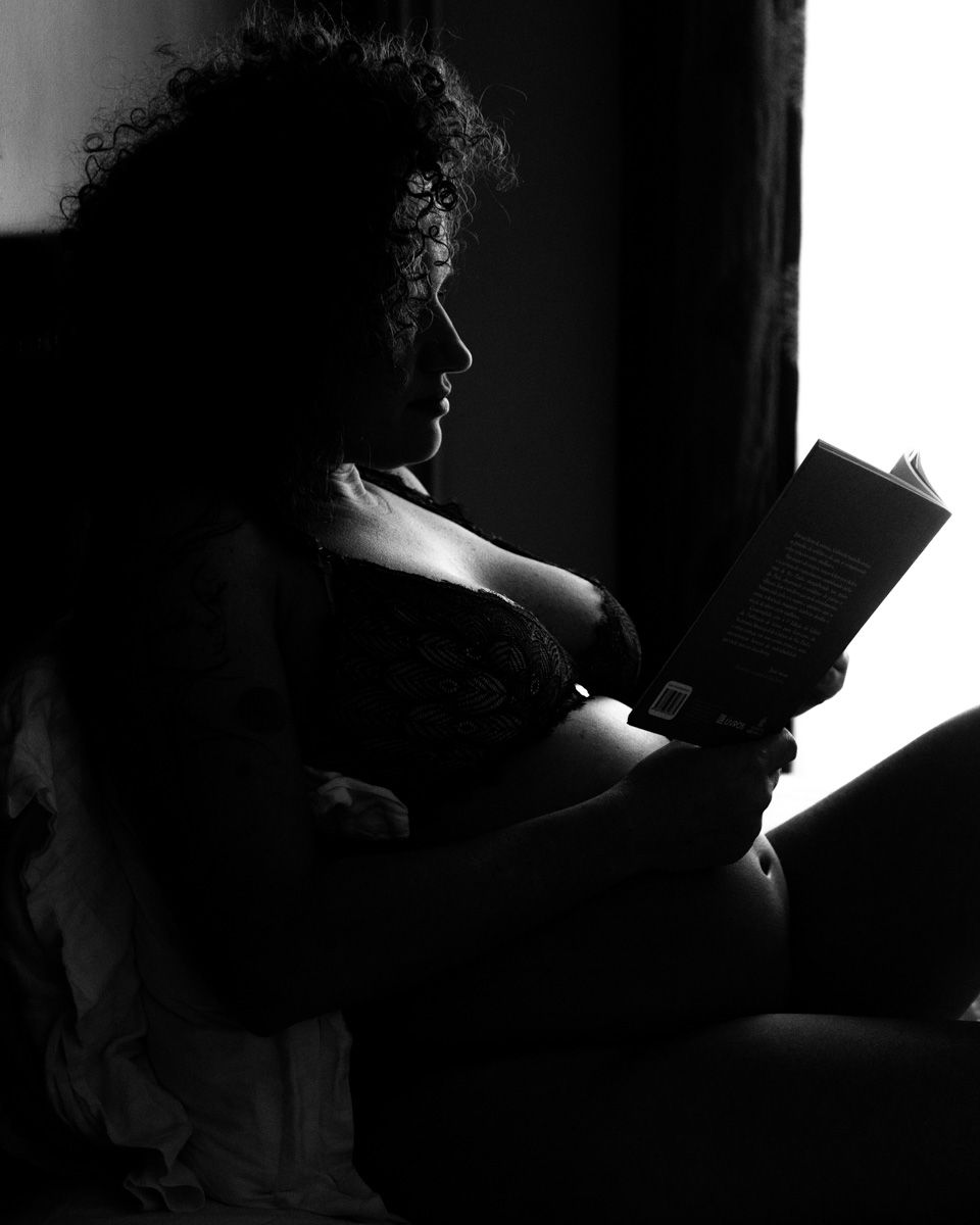 Black and white silhouette of pregnant woman reading a book by window light.