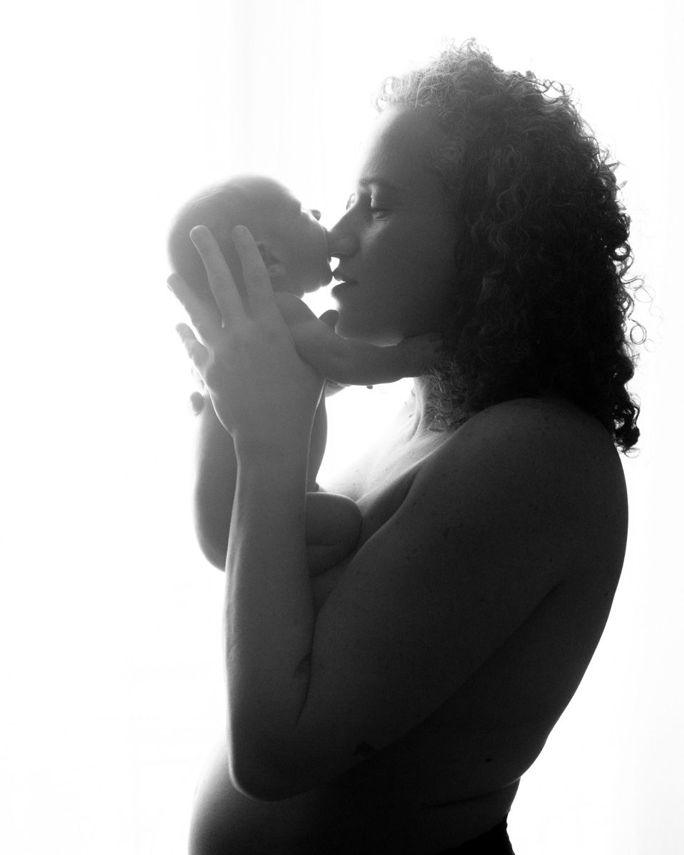 A silhouetted figure in a backlit black and white photo holds their baby near their face in a contemplative pose.