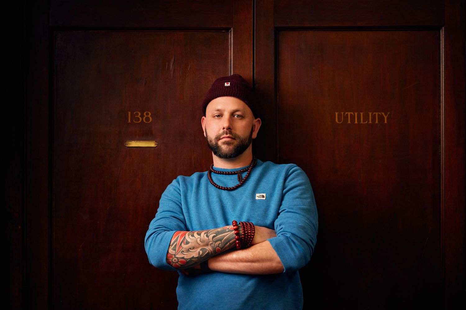 A person in blue sweatshirt with tattoos standing against wooden doors marked with utility signs.