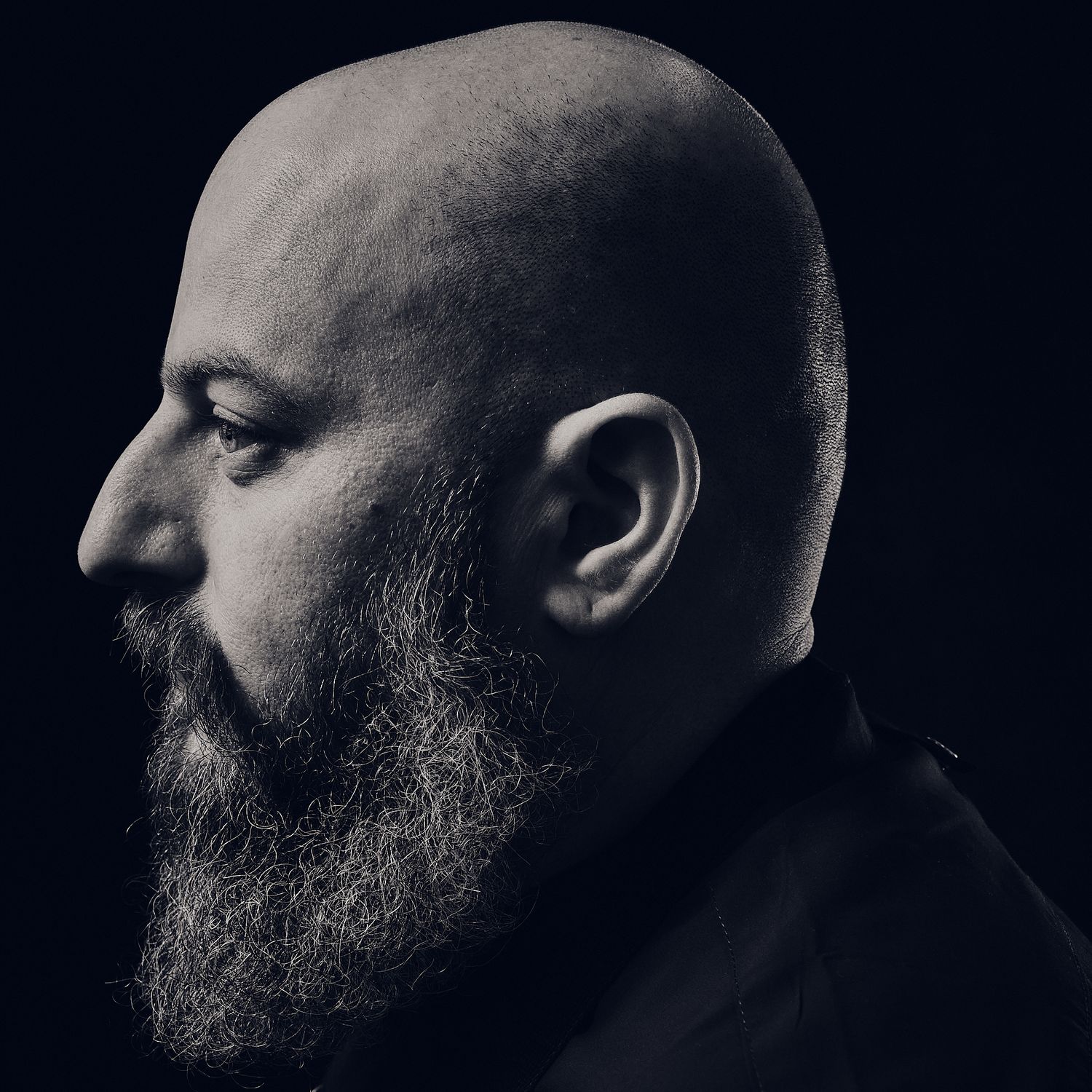 Side profile portrait of a person with a bald head and full beard against dark background.