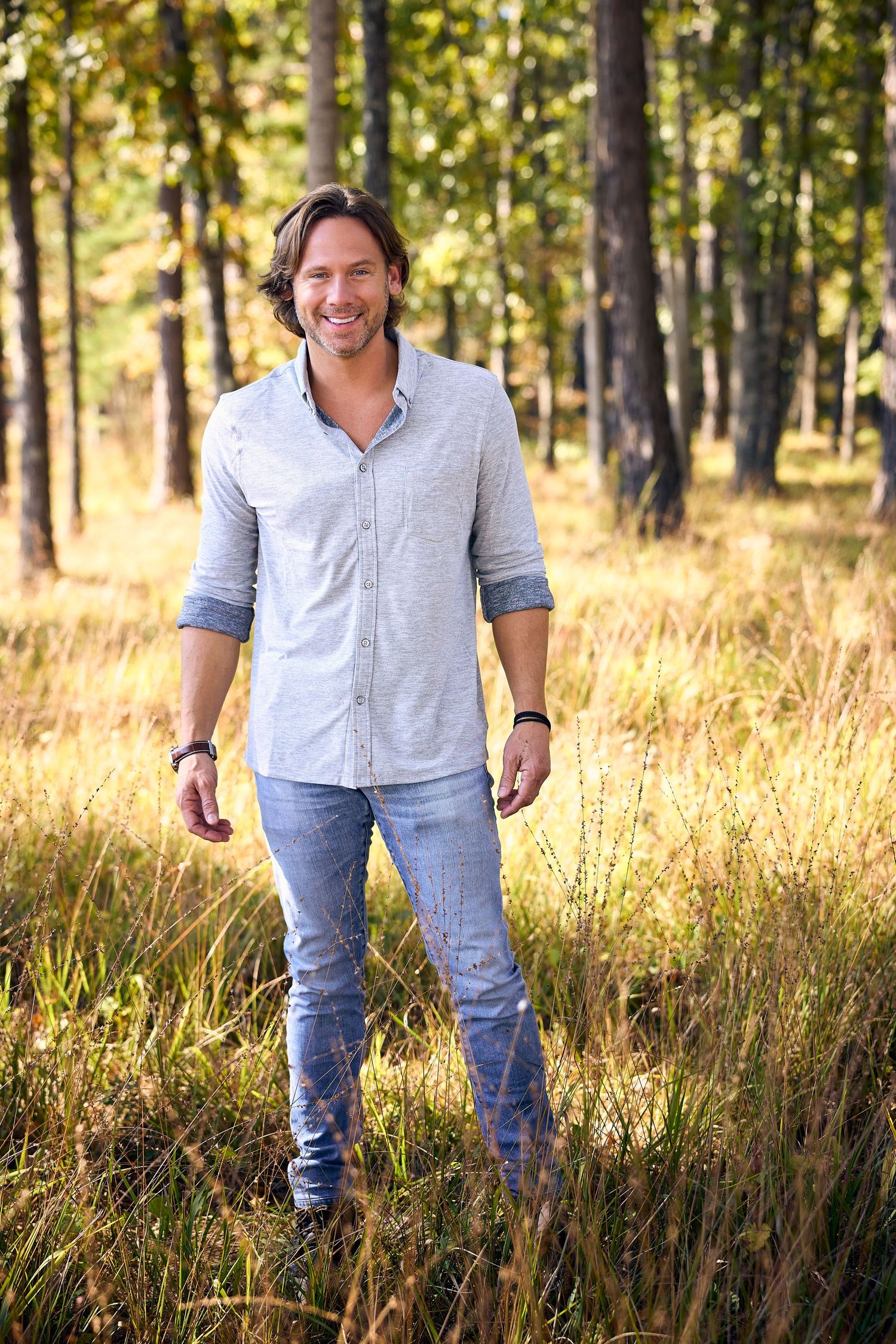 A person in casual attire standing in a sunlit forest with tall pine trees.