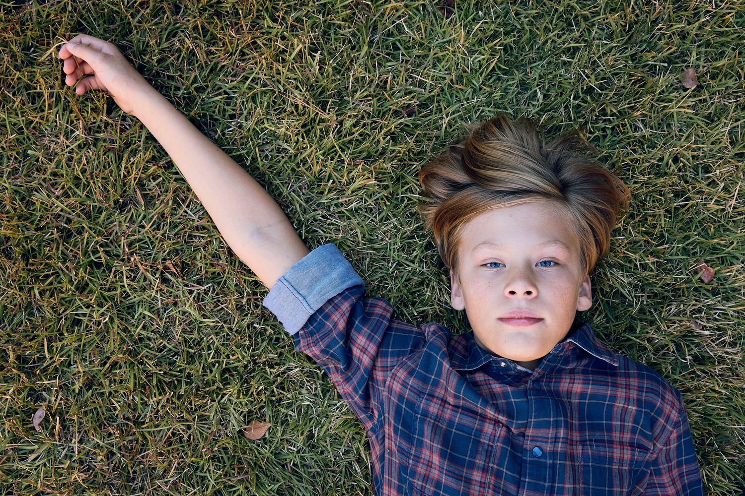 Young boy lying on grass with arms outstretched wearing a plaid shirt.
