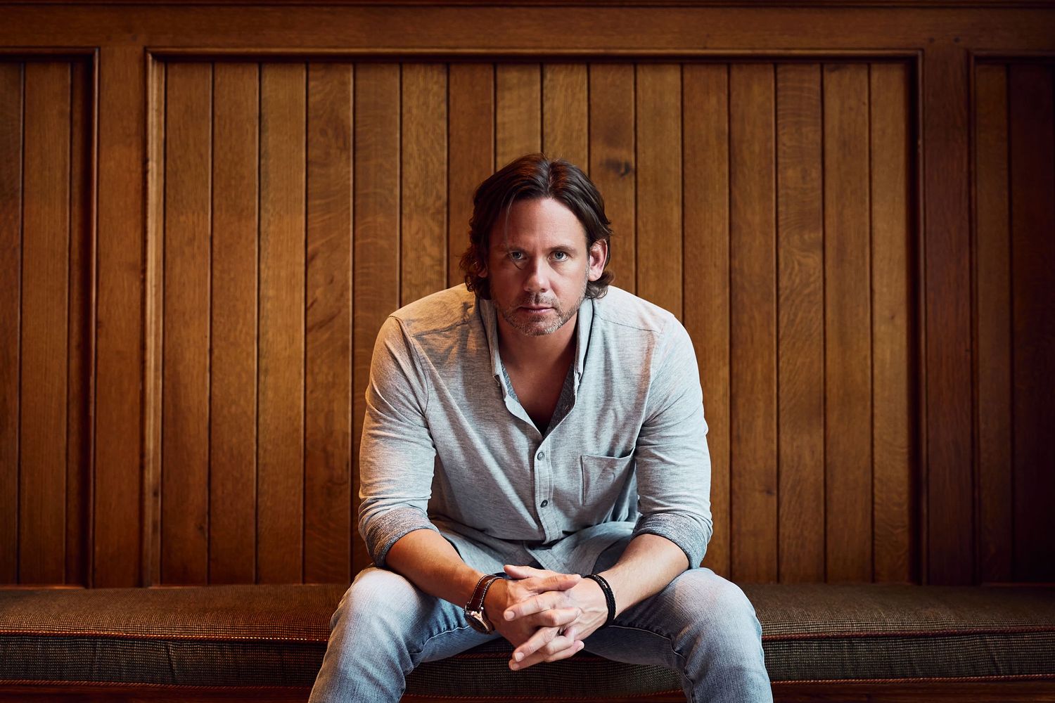 A casual portrait of a person seated on a couch against wood paneling wearing a light button-down shirt.