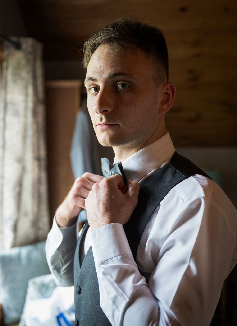 Man in white dress shirt and black vest adjusts his tie while looking in mirror in dim lit room.