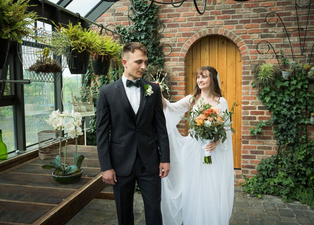 Wedding couple poses by brick wall with arched wooden door and hanging plants in garden setting.