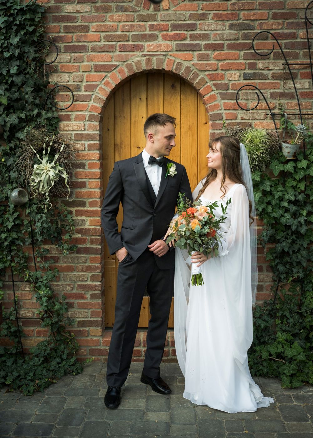 Wedding couple poses against rustic brick wall with wooden door and climbing ivy decorations.