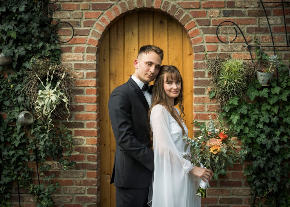 Elegant wedding portrait in front of rustic wooden door framed by ivy and brick archway.
